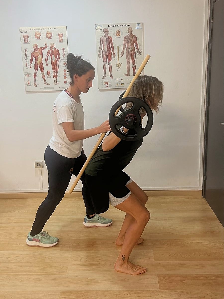 Una fisioterapeuta ajuda una dona a realitzar un exercici de força amb una barra i discs de pes en una sala de fisioteràpia.